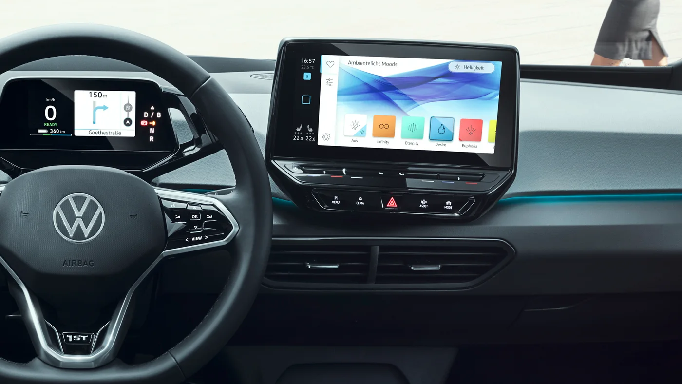 Volkswagen ID.3 console. Beneath the touchscreen is a flush capacitive strip for temperature and volume, and below that a row of flat touch icons. The steering wheel spokes carry smooth black haptic pads. Nothing on the dash moves when you press it.