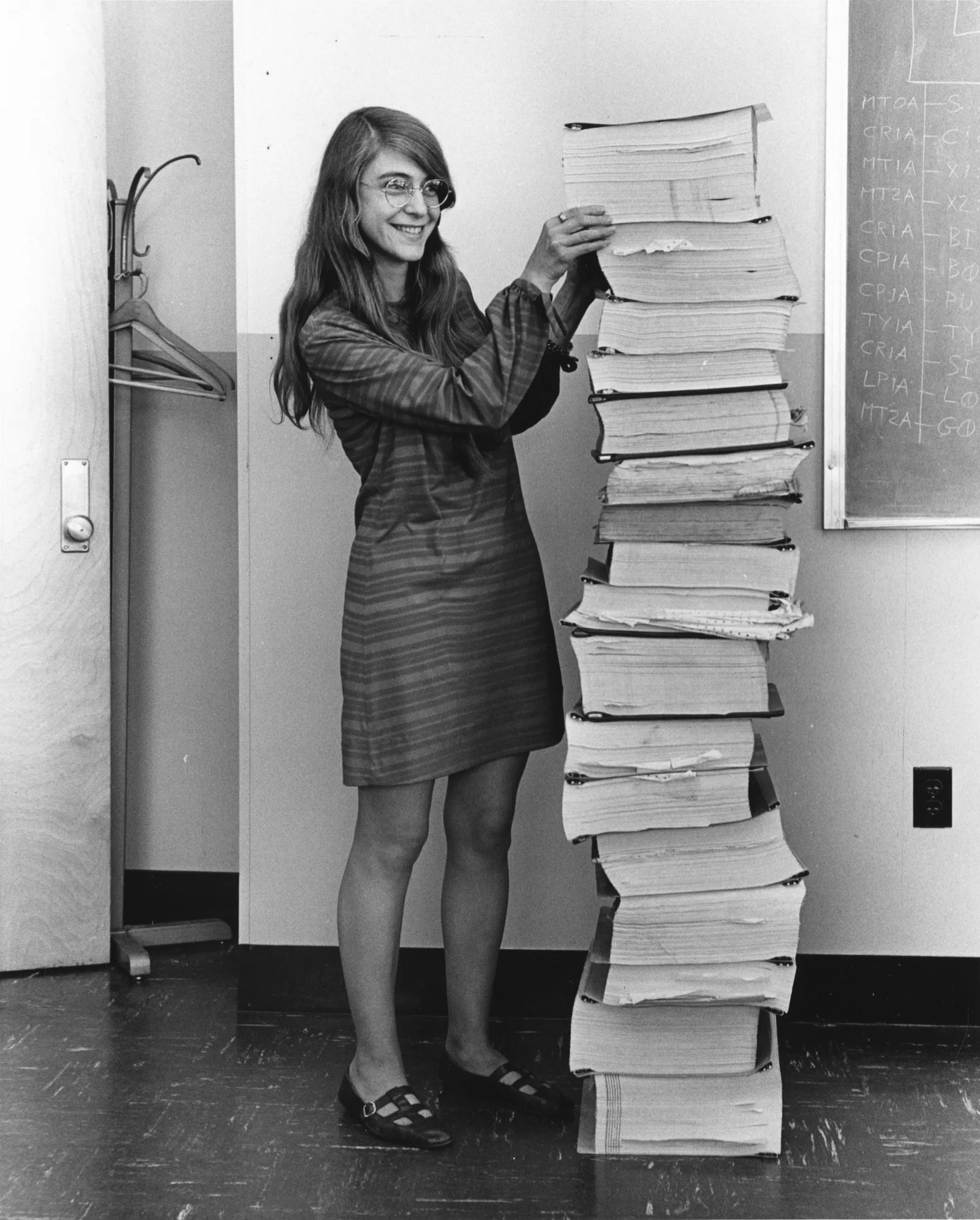 Margaret Hamilton beside a stack of printed Apollo flight-software listings that rises well above her head.