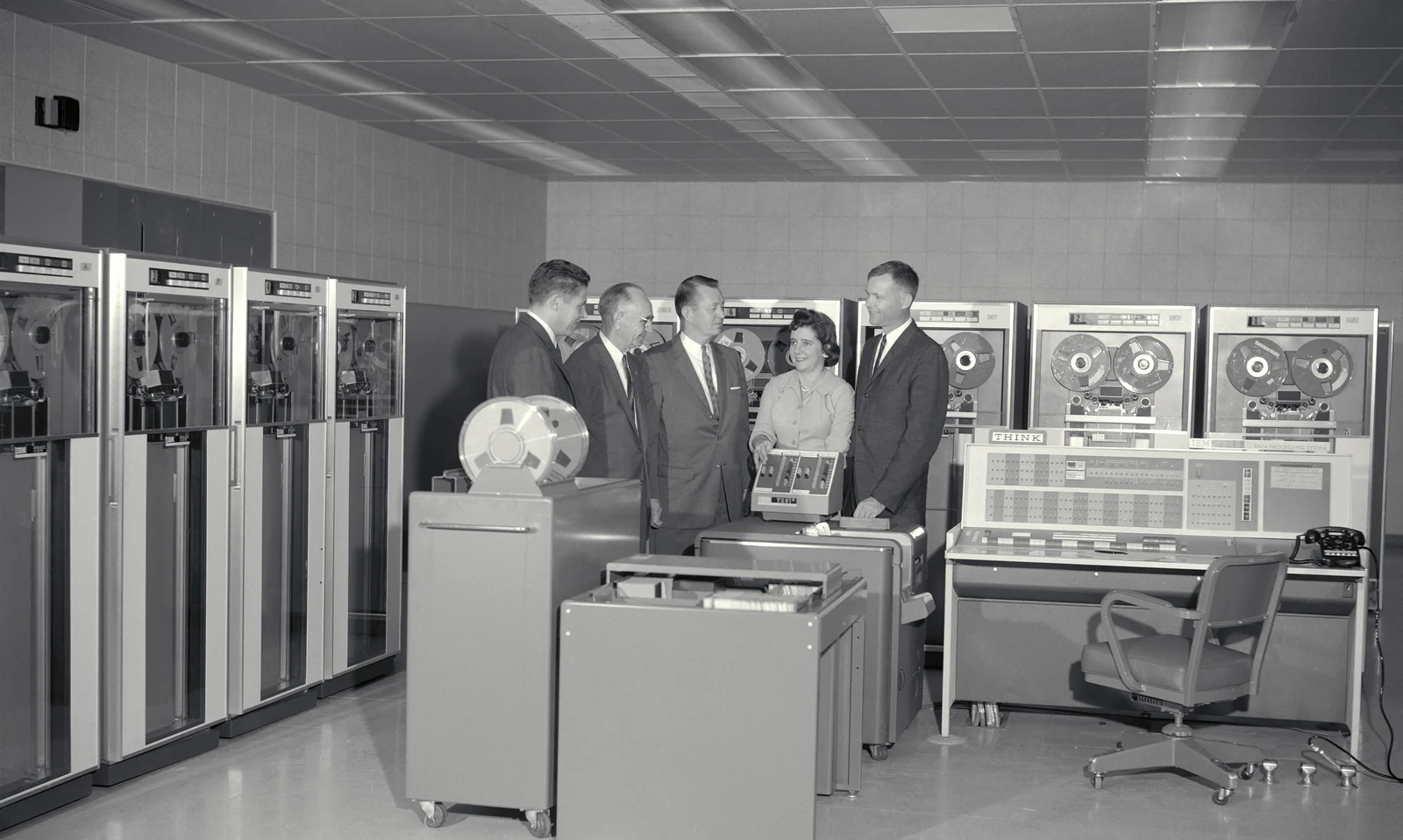 Five people stand among tall tape drives and a control console in a 1961 NASA computer room.